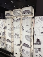 A series of illuminated cuboid structures featuring intricate black sketches of traditional Asian architecture and cultural symbols. The designs are arranged in a collage style on a light background, creating a striking contrast. The setting appears modern with a ceiling grid pattern visible above.