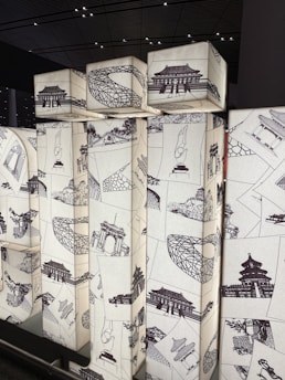 A series of illuminated cuboid structures featuring intricate black sketches of traditional Asian architecture and cultural symbols. The designs are arranged in a collage style on a light background, creating a striking contrast. The setting appears modern with a ceiling grid pattern visible above.