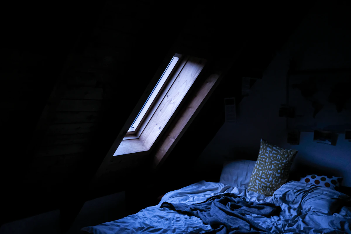Habitación de ático en penumbra, con luz entrando por una claraboya sobre una cama deshecha