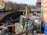 The image features a construction site by a river with metal barriers and various equipment, including pumps and pipes. In the background, there are buildings and leafless trees under a clear sky. Graffiti is visible on some barriers, and there is signage related to the construction work.