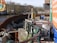 The image features a construction site by a river with metal barriers and various equipment, including pumps and pipes. In the background, there are buildings and leafless trees under a clear sky. Graffiti is visible on some barriers, and there is signage related to the construction work.