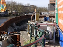 The image features a construction site by a river with metal barriers and various equipment, including pumps and pipes. In the background, there are buildings and leafless trees under a clear sky. Graffiti is visible on some barriers, and there is signage related to the construction work.
