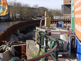 The image features a construction site by a river with metal barriers and various equipment, including pumps and pipes. In the background, there are buildings and leafless trees under a clear sky. Graffiti is visible on some barriers, and there is signage related to the construction work.