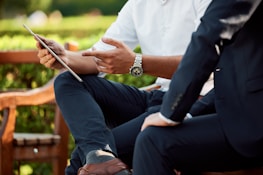 Two individuals engaged in a discussion while seated outdoors, with one holding a tablet and gesturing with a hand. They are dressed in business attire, and there is a lush green background.