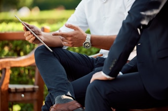 Two individuals engaged in a discussion while seated outdoors, with one holding a tablet and gesturing with a hand. They are dressed in business attire, and there is a lush green background.