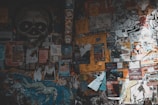 A wall covered with numerous posters, stickers, and graffiti. The posters are of various sizes, colors, and themes, with some promoting events, bands, and advertisements. A large black and white skull graphic is prominent on the left side. There are also drawings and colorful illustrations scattered throughout the wall.