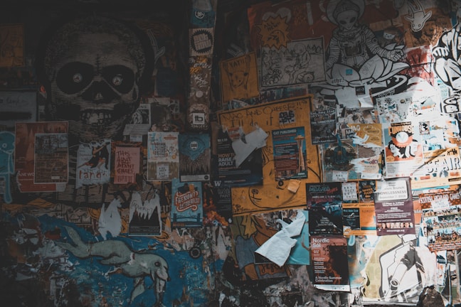 A wall covered with numerous posters, stickers, and graffiti. The posters are of various sizes, colors, and themes, with some promoting events, bands, and advertisements. A large black and white skull graphic is prominent on the left side. There are also drawings and colorful illustrations scattered throughout the wall.