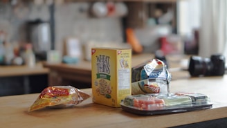 A countertop featuring an assortment of snacks including a box of Wheat Thins, a bag of chips, a package of tortillas, and a tray of sushi. The background shows a blurred kitchen setting with various items on shelves.