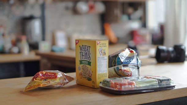 A countertop featuring an assortment of snacks including a box of Wheat Thins, a bag of chips, a package of tortillas, and a tray of sushi. The background shows a blurred kitchen setting with various items on shelves.