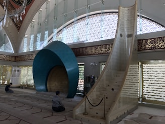 A spacious interior of a mosque featuring an intricately designed mihrab with a large blue arch. Arabic calligraphy adorns the walls, and natural light filters through patterned windows. Two people are seated on the carpet, appearing to be in prayer.
