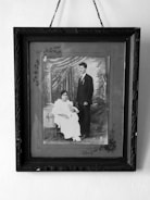 A sepia-toned framed photograph depicts a seated woman in a long white dress holding a baby, alongside a man in a dark suit standing beside them. The backdrop includes ornate drapery and classic studio props.