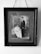 A sepia-toned framed photograph depicts a seated woman in a long white dress holding a baby, alongside a man in a dark suit standing beside them. The backdrop includes ornate drapery and classic studio props.