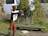 A metal sculpture of a humanoid figure is positioned in a grassy area, holding what appears to be a can or object. The sculpture is rusted and stands near a wooden bench. There is foliage in the background including yellow flowering plants. A white bus is parked behind the greenery, and a circular black sign with white text is visible near the sculpture.