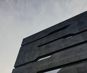 A modern architectural building with a minimalist design. The structure features dark, sleek lines and geometric shapes against a cloudy sky backdrop.