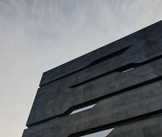 A modern architectural building with a minimalist design. The structure features dark, sleek lines and geometric shapes against a cloudy sky backdrop.