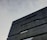 A modern architectural building with a minimalist design. The structure features dark, sleek lines and geometric shapes against a cloudy sky backdrop.