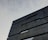 A modern architectural building with a minimalist design. The structure features dark, sleek lines and geometric shapes against a cloudy sky backdrop.