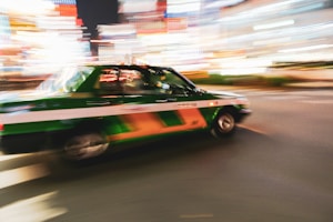 A green and white taxi is captured in motion against a backdrop of vibrant city lights at night, conveying a sense of speed and energy.