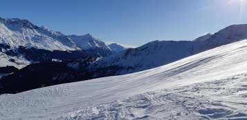 A picturesque mountain landscape covered in snow with clear blue skies. The image captures a series of snow-covered peaks and ridges, creating a serene and tranquil winter scene.