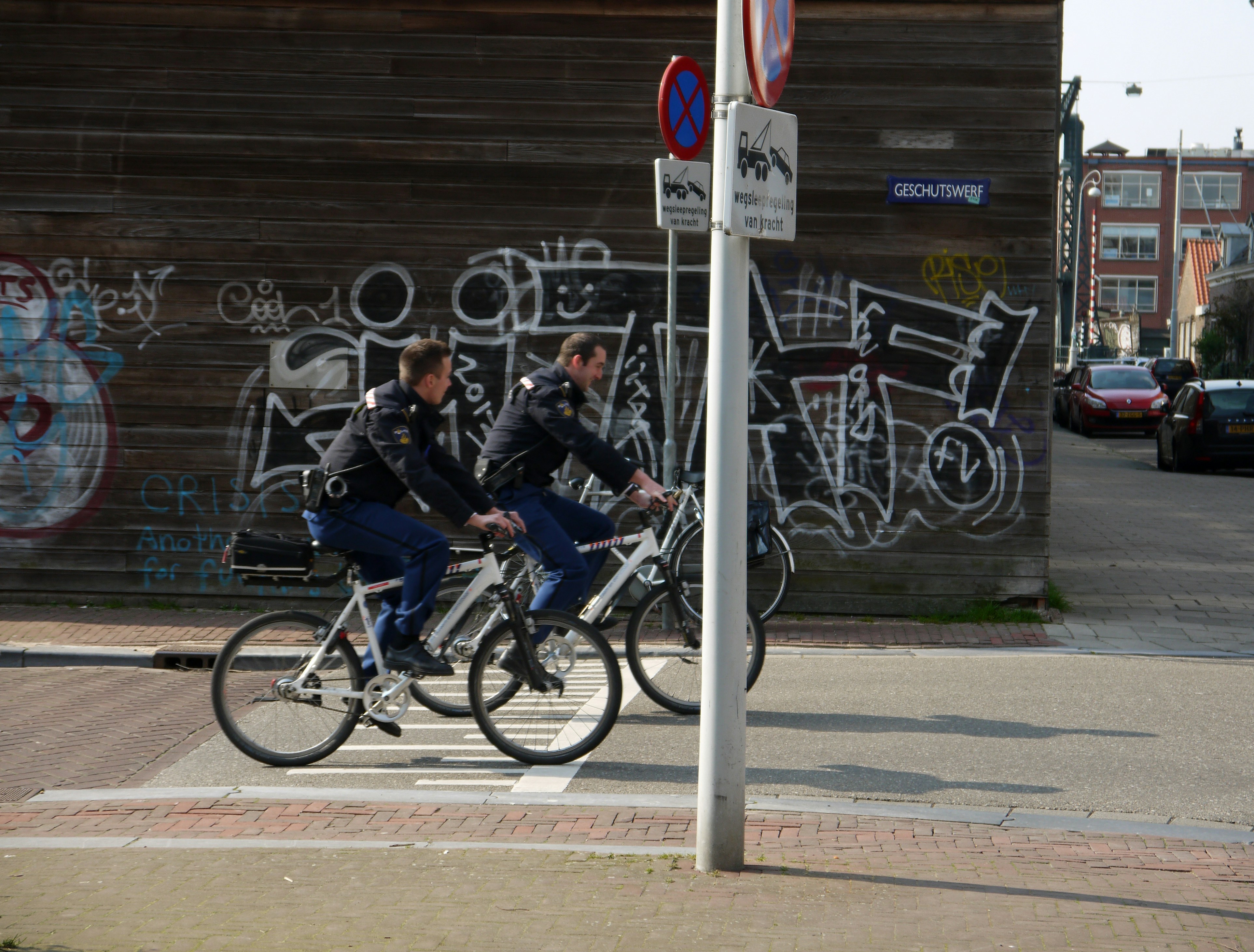 two men riding bikes on road at the city during day, Free photo of Amsterdam city - a picture of two cops on the bike in the street in the sunlight of early Spring. Large graffiti tags on a wooden wall, as background of the uniforms and policemen. Sunny street in late March 2014. Urban photography of The Netherlands by Fons Heijnsbroek, March 2014. Dutch version: Foto Amsterdam - Twee agenten op de fiets met op de achtergrond in de zonnige straat een houten muur met grote graffiti tags, gespoten in zwart-wit; alles in het licht van vroege lente. Het is in het zonlicht van Maart 2014.