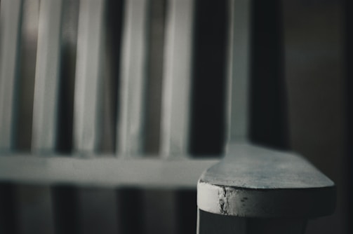 Close-up of a worn wooden chair armrest with a blurred background. The focus is on the texture and wear of the armrest, creating a sense of age and character.