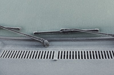 A close-up view of a car windshield with frost covering the glass surface. The windshield wipers are visible, covered in a layer of frost. Water droplets are scattered across the frosty surface, adding texture.