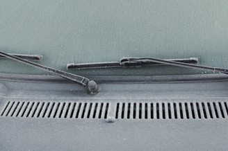 A close-up view of a car windshield with frost covering the glass surface. The windshield wipers are visible, covered in a layer of frost. Water droplets are scattered across the frosty surface, adding texture.