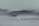 A close-up view of a car windshield with frost covering the glass surface. The windshield wipers are visible, covered in a layer of frost. Water droplets are scattered across the frosty surface, adding texture.