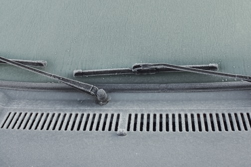 A close-up view of a car windshield with frost covering the glass surface. The windshield wipers are visible, covered in a layer of frost. Water droplets are scattered across the frosty surface, adding texture.