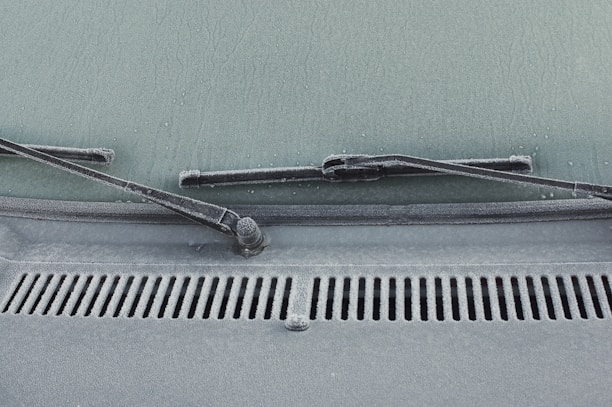 A close-up view of a car windshield with frost covering the glass surface. The windshield wipers are visible, covered in a layer of frost. Water droplets are scattered across the frosty surface, adding texture.