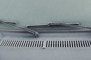 A close-up view of a car windshield with frost covering the glass surface. The windshield wipers are visible, covered in a layer of frost. Water droplets are scattered across the frosty surface, adding texture.