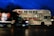 A double-decker bus featuring an advertisement for a vodka brand, with the text 'GREAT GETAWAY CARS' and a large image of a vodka bottle. The bus is stationary on a street at night, and a black van is passing in front of it. The background is dark with silhouetted trees and a deep blue sky.