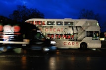 A double-decker bus featuring an advertisement for a vodka brand, with the text 'GREAT GETAWAY CARS' and a large image of a vodka bottle. The bus is stationary on a street at night, and a black van is passing in front of it. The background is dark with silhouetted trees and a deep blue sky.