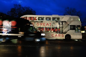 A double-decker bus featuring an advertisement for a vodka brand, with the text 'GREAT GETAWAY CARS' and a large image of a vodka bottle. The bus is stationary on a street at night, and a black van is passing in front of it. The background is dark with silhouetted trees and a deep blue sky.
