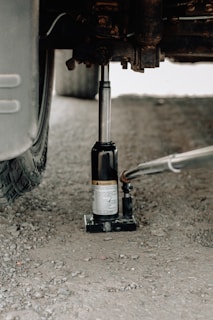 A hydraulic car jack is being used to lift a vehicle on a gravel surface. The focus is on the metallic jack under the car, with a visible tire on the left side. The ground is uneven and consists of small stones and dirt.