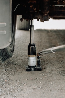 A hydraulic car jack is being used to lift a vehicle on a gravel surface. The focus is on the metallic jack under the car, with a visible tire on the left side. The ground is uneven and consists of small stones and dirt.