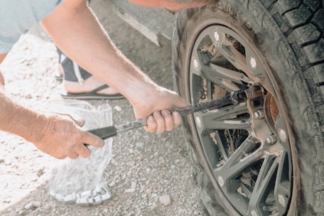 A person is using a lug wrench to tighten or loosen the bolts on a car tire. The setting appears to be outdoors, possibly on a gravel surface. The tire and wheel are dusty, suggesting off-road use. The person is wearing sandals.