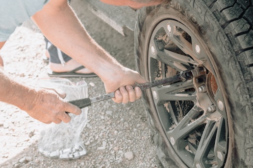 A person is using a lug wrench to tighten or loosen the bolts on a car tire. The setting appears to be outdoors, possibly on a gravel surface. The tire and wheel are dusty, suggesting off-road use. The person is wearing sandals.