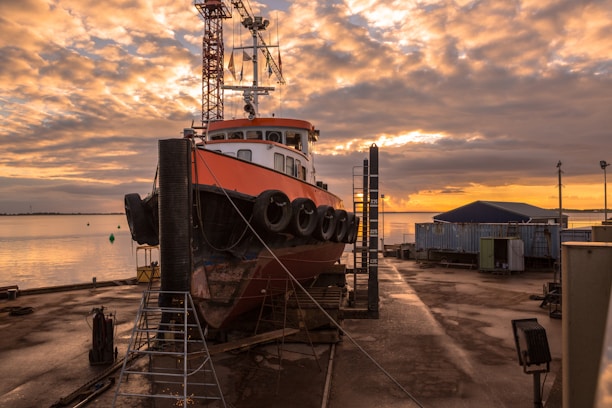 A large boat is docked for maintenance on a shipyard beside a calm body of water during sunset. The sky is filled with dramatic clouds illuminated in shades of orange and yellow. The scene includes industrial elements such as storage containers, ladders, and cranes surrounding the ship.