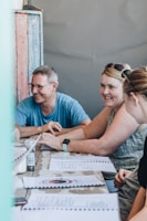 A group of people sitting at a table engaged in a conversation. Two individuals are smiling, and there are menus on the table. A wooden wall with a distressed paint finish is in the background, and there is a relaxed and casual atmosphere.