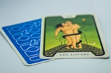A tarot card titled 'The Lovers' with an illustration of two figures embracing on a green background. The card is placed on top of another card with a blue geometric pattern.