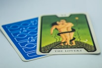 A tarot card titled 'The Lovers' with an illustration of two figures embracing on a green background. The card is placed on top of another card with a blue geometric pattern.