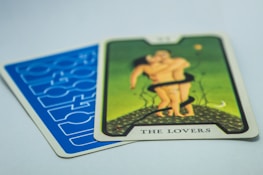 A tarot card titled 'The Lovers' with an illustration of two figures embracing on a green background. The card is placed on top of another card with a blue geometric pattern.
