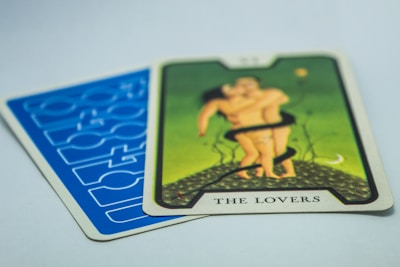A tarot card titled 'The Lovers' with an illustration of two figures embracing on a green background. The card is placed on top of another card with a blue geometric pattern.