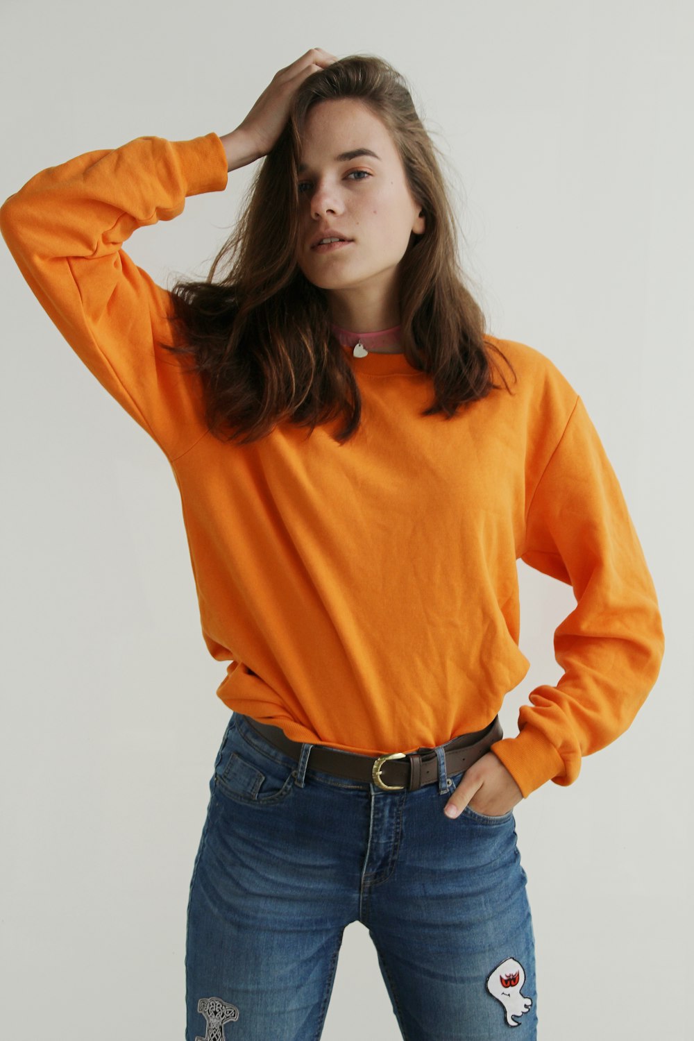 オレンジ色のクルーネックのスウェットシャツを着て、右手を頭に置いて立っている女性