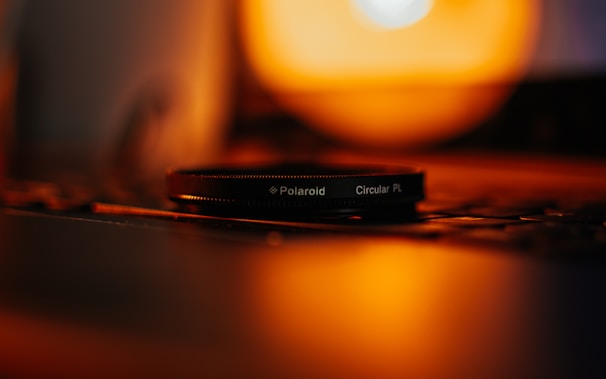 A camera lens filter labeled 'Polaroid Circular PL' is placed on a surface with a blurred background of warm, orange lighting. The lighting creates a soft and cozy ambiance, reflecting off the surface.