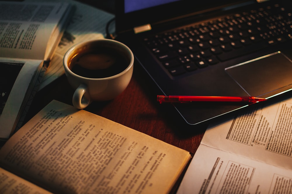 cup of coffee in between of open book and black laptop