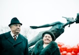 An elderly man and woman stand in front of a modern bronze sculpture depicting two figures in an abstract embrace. The man wears a formal dark coat and hat, while the woman wears a fur-collared coat and a beret. Behind them, numerous red balloons are visible, creating a vivid contrast against the sculpture's dark tones.