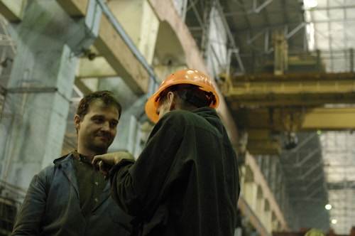 Two individuals are engaged in conversation inside an industrial facility. One person is wearing a work jacket, and the other is wearing a hard hat, suggesting they are in a professional setting related to construction or manufacturing. The large industrial cranes and high ceilings visible in the background indicate a factory or warehouse environment.