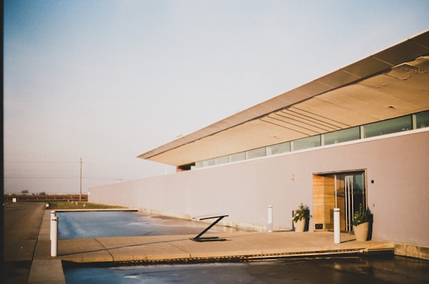 A modern, minimalist building with clean lines and a flat roof is presented under a clear sky. The structure has large glass windows and light-colored walls. There is a concrete patio with a reflective water feature in front, along with several potted plants. The setting appears tranquil and spacious, hinting at a suburban or rural location.